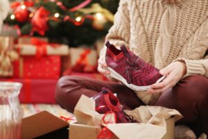 A woman in a beige cable knit sweater is unwrapping her gifts; a pair of red running shoes.