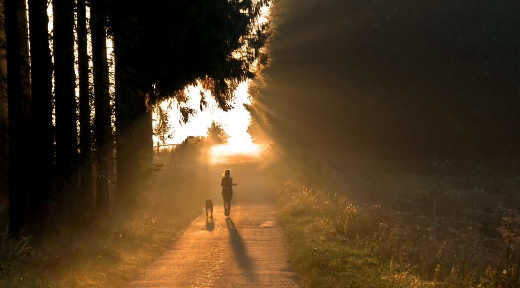 A woman with a dog is running on a foggy morning down a country lane, managing her chronic illness with exercise.