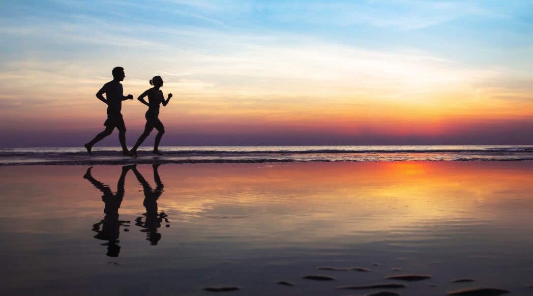 Silhouettes of two runners, a woman and a man, running on a beach by the ocean after sunset, enjoying the health benefits of running